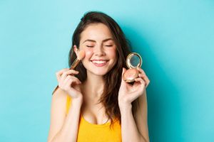 beauty-close-up-beautiful-young-woman-close-eyes-smile-while-applying-blushes-cheeks-with-make-up-brush-standing-happy-against-blue-background beauty-close-up-beautiful-young-woman-close-eyes-smile-while-applying-blushes-cheeks-with-make-up-brush-standing-happy-against-blue-background